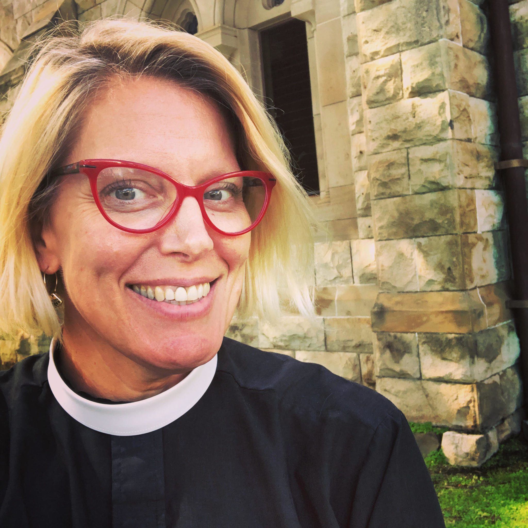 Woman, Heather Patton-Graham, Staff Consultant, in Roman collar, standing in front of a church, smiling